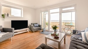 A modern living room with a flat-screen TV, comfortable seating, a coffee table, and large windows offering a view of the beach. - 3 Bed in Rustington (oc-p45430) (Rustington)