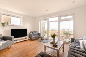 Ein modernes Wohnzimmer mit Fernseher, bequemen Sitzmöglichkeiten, einem Couchtisch und großen Fenstern mit Blick auf den Strand.