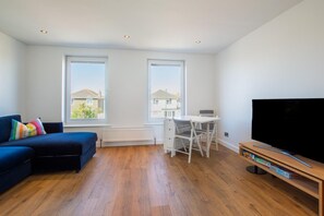 A modern living room with a blue sofa, a dining table with chairs, a flat-screen TV, and large windows offering a view of the neighborhood.
