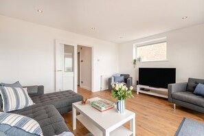 A modern living room with a grey sectional sofa, a white coffee table with a book and flowers, a grey armchair, and a flat-screen TV against a brick wall. - 3 Bed in Rustington (oc-p45430) (Rustington)