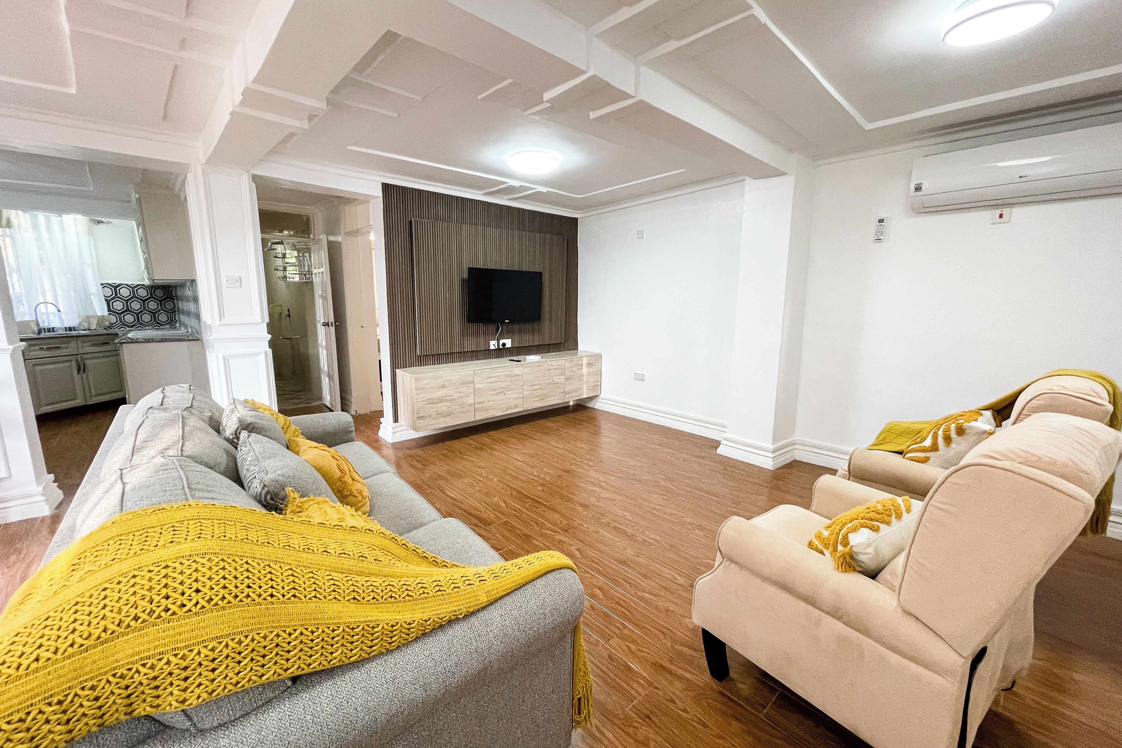 Un salon moderne avec un ensemble de canapés, un téléviseur fixé au mur et un coin cuisine à l’arrière-plan.