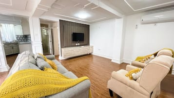 Un salon moderne avec un ensemble de canapés, un téléviseur fixé au mur et un coin cuisine à l’arrière-plan.