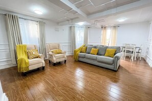 Un salon comprenant un canapé, deux fauteuils, une table à manger avec des chaises et un plafond doté d’un éclairage encastré.