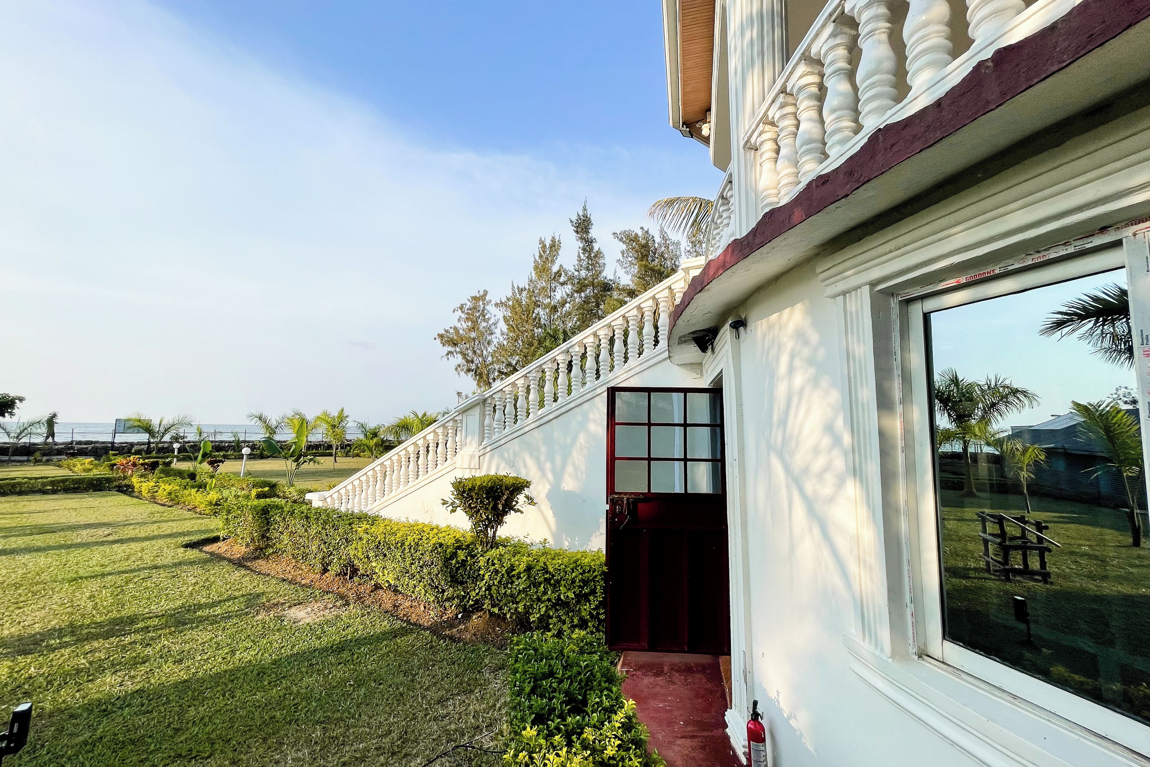 Un bâtiment à l’extérieur blanc, avec une porte d’entrée rouge et une porte vitrée. On y trouve une pelouse bien entretenue, agrémentée de diverses plantes, et un ciel dégagé.