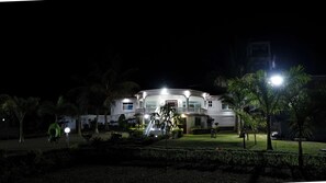 A large, white building with multiple windows and a balcony, surrounded by well-maintained lawns and palm trees.