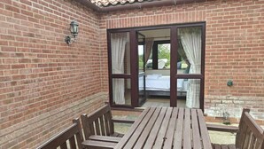 A patio with a wooden table and benches, a brick wall, and a sliding glass door leading to a bed. - 2 Bed in Acle (oc-s45732) (Acle)
