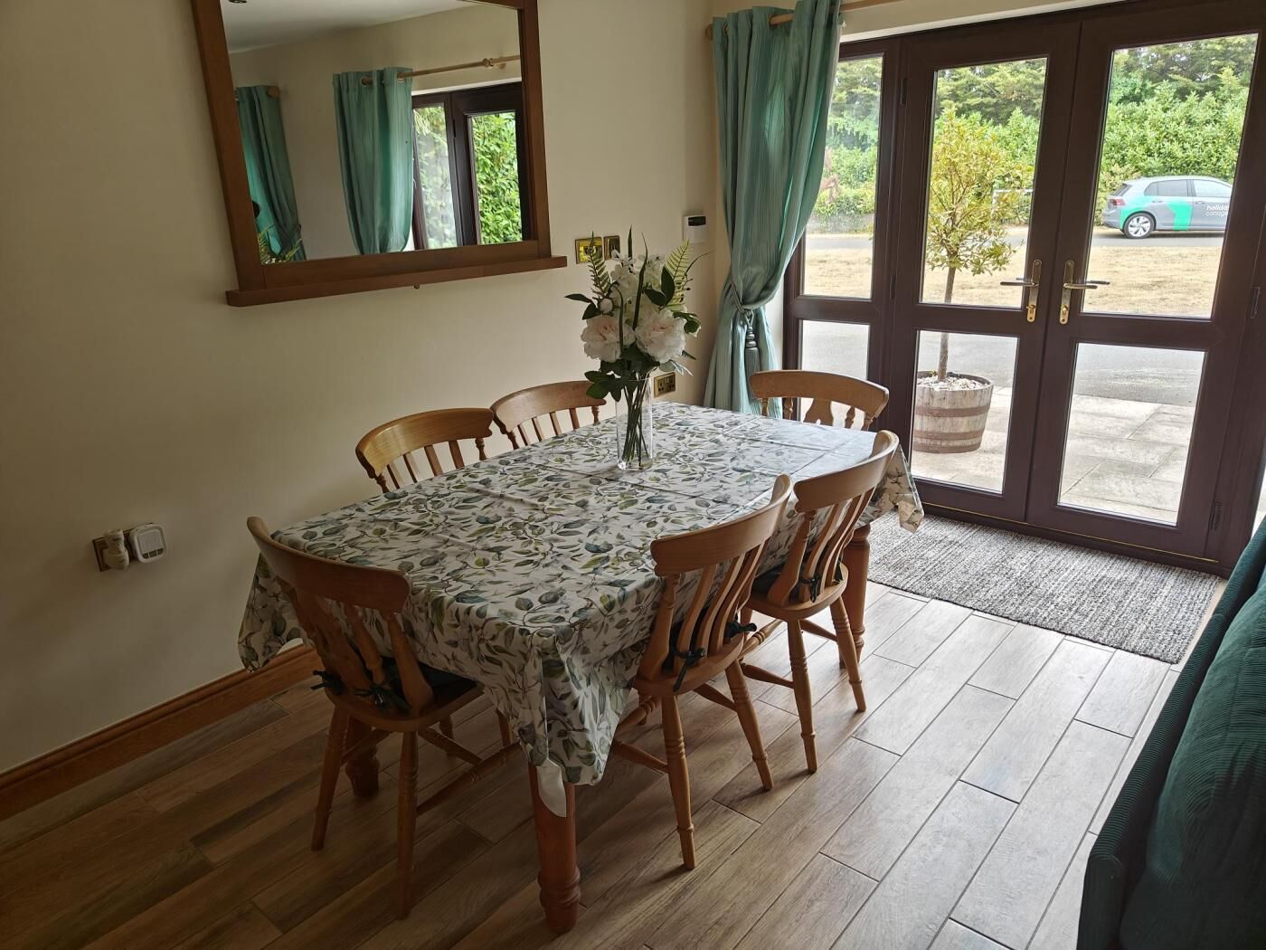 A dining area with a table set for four, wooden chairs, a vase with flowers, a mirror, and a view of the outside through a glass door.