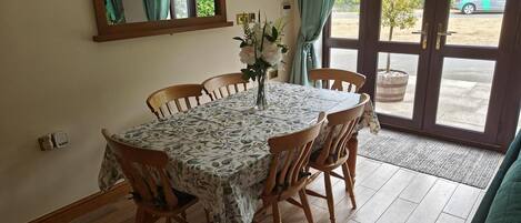 A dining area with a table set for four, wooden chairs, a vase with flowers, a mirror, and a view of the outside through a glass door.