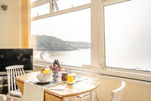 A dining area with a table set for breakfast, overlooking a beach with cliffs. - 1 Bed in Caswell Bay (oc-h45734) (Bishopston)
