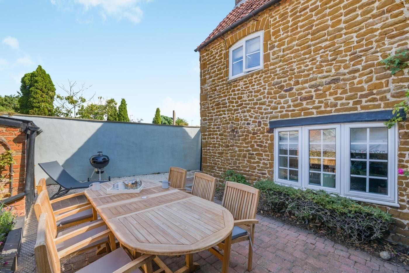 A stone house with a patio area featuring a wooden table and chairs, a barbecue grill, and a window.