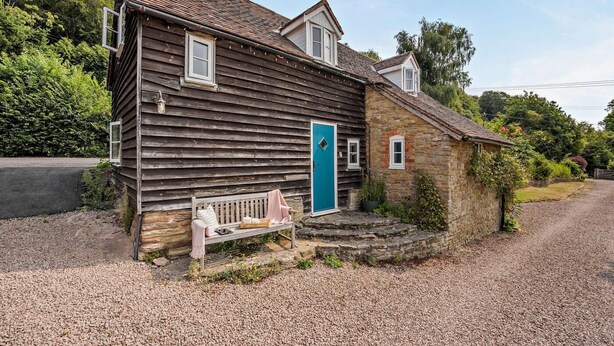 A wooden house with a blue door, a bench outside, and a stone building nearby. - 2 Bed in Cradley (oc-s45721) (Cradley and Storridge)