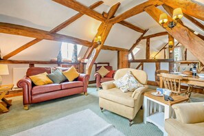 Ein Loft-Zimmer mit freiliegenden Holzbalken, einem roten Ledersofa, einem beigen Sessel und einem Esstisch mit einer Vase und Brot.