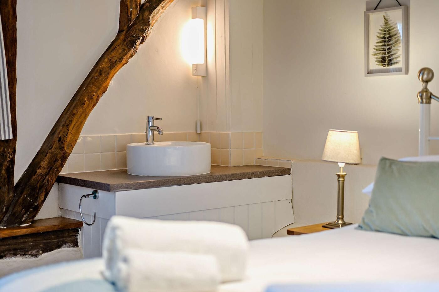 Ein Hotelzimmer mit einem runden weißen Waschbecken, einer Nachttischlampe, einer gerahmten Pflanze und einem Bett mit einem weißen Kopfkissen.