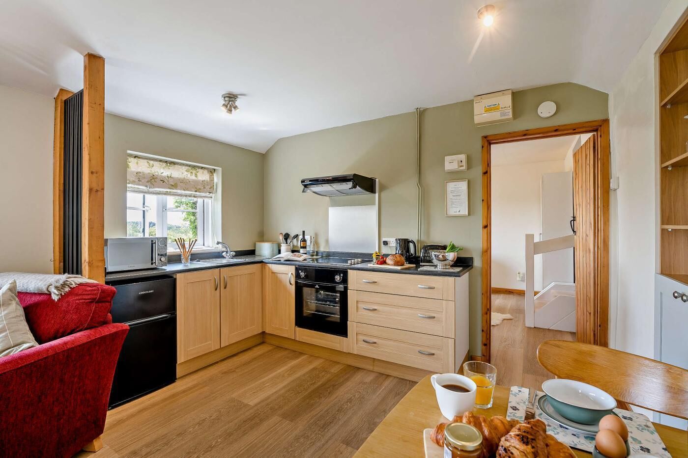 A compact kitchen with a microwave, stove, and cabinets, a dining table with breakfast items, and a red chair.