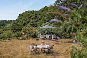 An outdoor dining area with a table and chairs, surrounded by lush greenery and a hill in the background. - 1 Bed in Cradley (oc-s45723) (Cradley and Storridge)