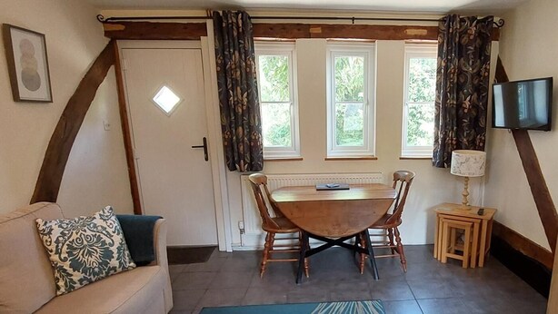 A cozy room with a wooden dining table, two chairs, a sofa with a patterned pillow, a small side table with a lamp, and a TV mounted on the wall. - 1 Bed in Cradley (oc-s45723) (Cradley and Storridge)
