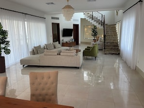 Un amplio salón con un sofá modular grande, un sillón verde y una mesa de comedor. Una escalera con pared de piedra y barandilla de metal lleva a un nivel superior.