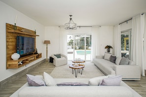 Ett rymligt vardagsrum med en stor TV, en trähylla, en soffa, ett soffbord och utsikt över en pool genom glasdörrarna.