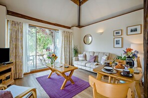 Ein möbliertes Wohnzimmer mit einer Couch, einem Esstisch mit Frühstücksgebäck, einem Fernseher und einem großen Fenster mit Vorhängen.