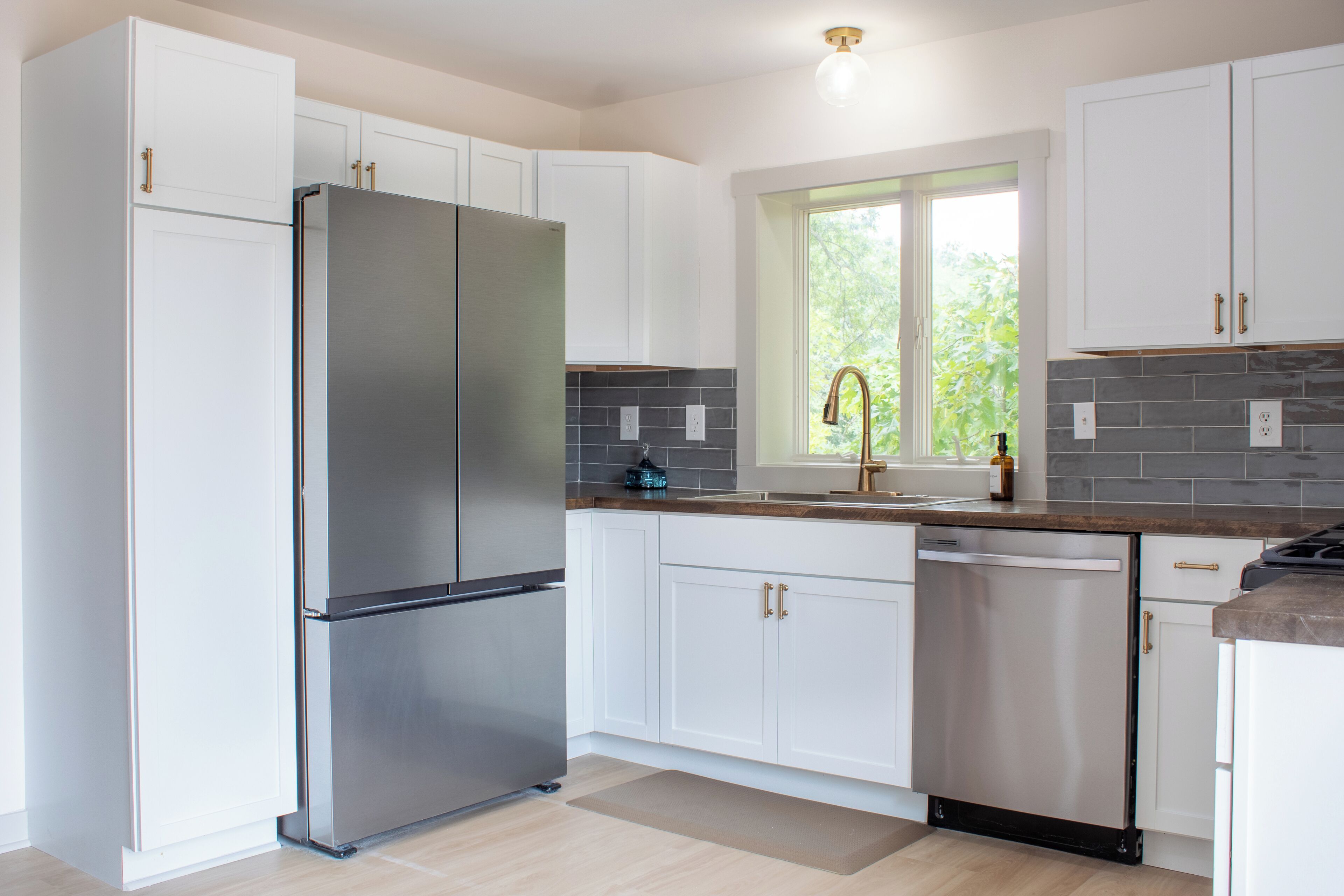 Cocina moderna con armarios blancos, refrigerador de acero inoxidable, lavavajillas y ventana con vista a follaje.