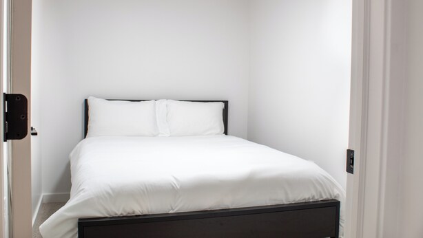 Habitación pequeña con una cama individual, una pared blanca lisa y una puerta a la izquierda.