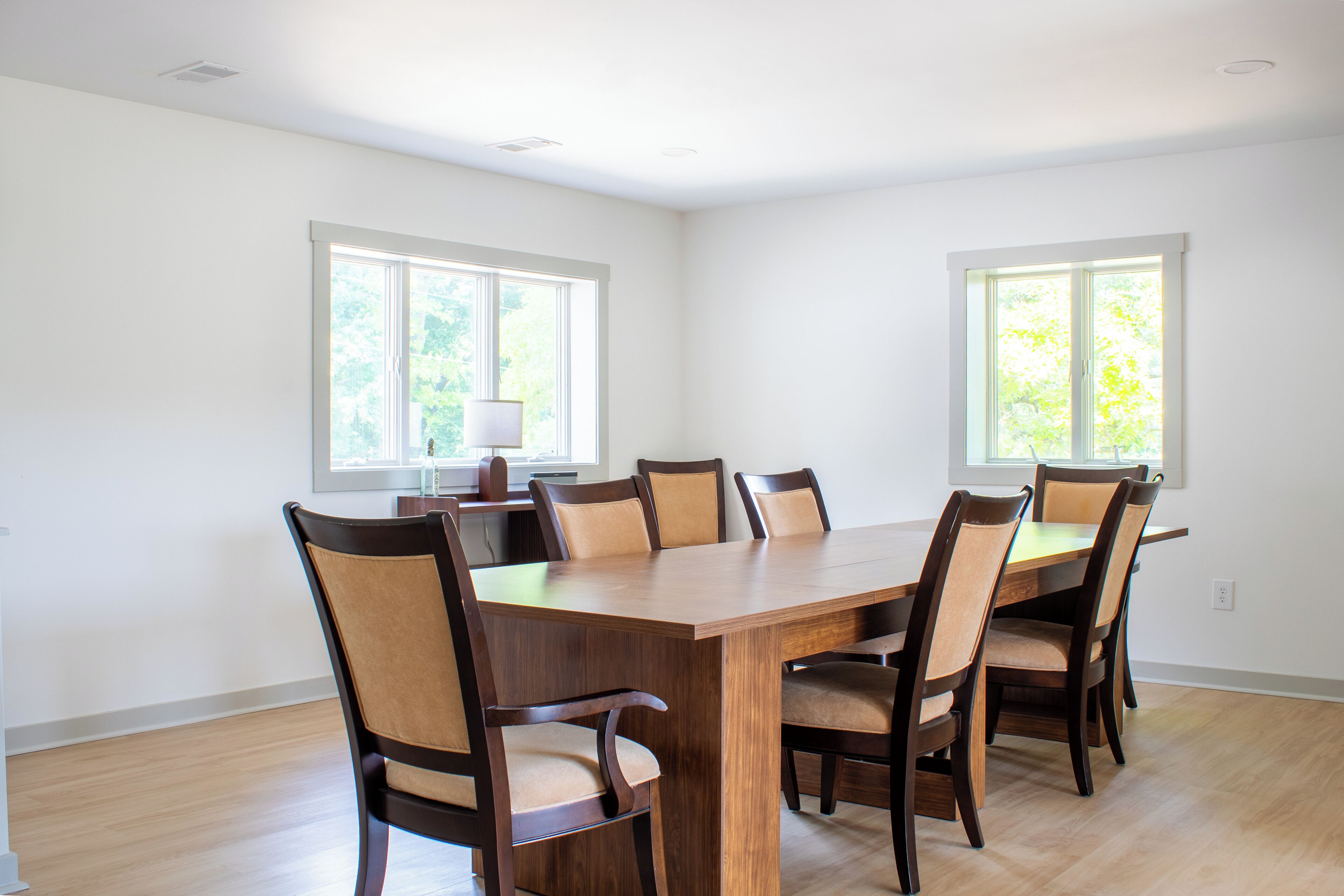 A spacious dining room with a large wooden table and six chairs, large windows with blinds, and a ceiling light fixture.