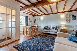 Ein Zimmer mit Holzboden, einer Couch, einem Teppich mit Muster, einem Esstisch mit Stühlen und einem Fenster mit Vorhängen.