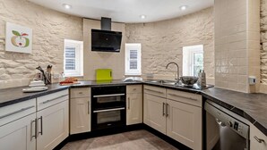 Een keuken met een achterwand van natuursteen, een inbouwoven, een vaatwasser en een raam met luxaflex.