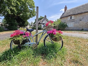 A bicycle with flower baskets parked on grass beside a road, with a stone house in the background. - 3 Bed in Owermoigne (oc-v42806) (Owermoigne)