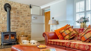 Una sala de estar acogedora con pared de piedra, chimenea, sofá de cuero con cojines estampados, una mesa de centro con tazas y un plato de comida, y una ventana con vistas.