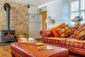 Ein gemütliches Wohnzimmer mit Steinwand, Kamin, Ledersofa mit gemusterten Kissen, Couchtisch mit Tassen und einem Teller Essen sowie ein Fenster mit Aussicht.