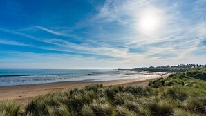 Un paisaje costero con una playa, dunas cubiertas de hierba y una línea de costa distante con edificios.