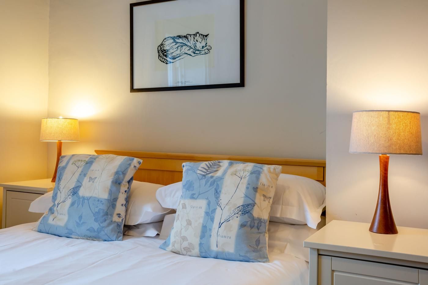 Habitación de hotel con cama, dos almohadas estampado botánico, mesita de noche con lámpara y un cuadro en la pared.