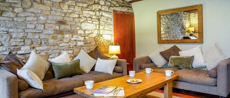 A cozy living room with stone walls, two sofas with cushions, a wooden coffee table with magazines, and a lamp.