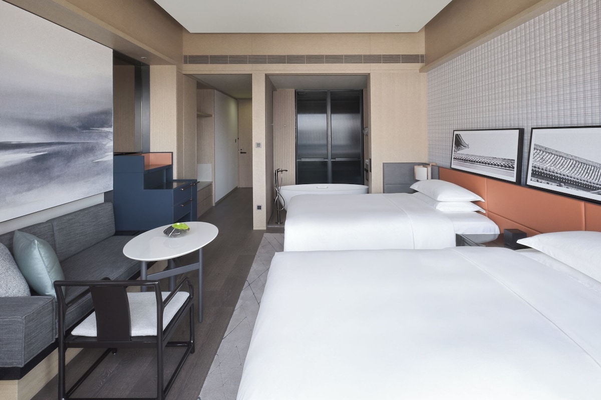 A modern hotel room with a large bed, a sofa, a small table, and a desk. There is a bathroom with a shower and a toilet. The room has a large window and a painting on the wall.