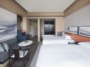 Habitación de hotel moderna con una cama grande, un sofá, una mesita y un escritorio. Cuenta con un baño con ducha e inodoro. La habitación tiene un ventanal grande y un cuadro en la pared.