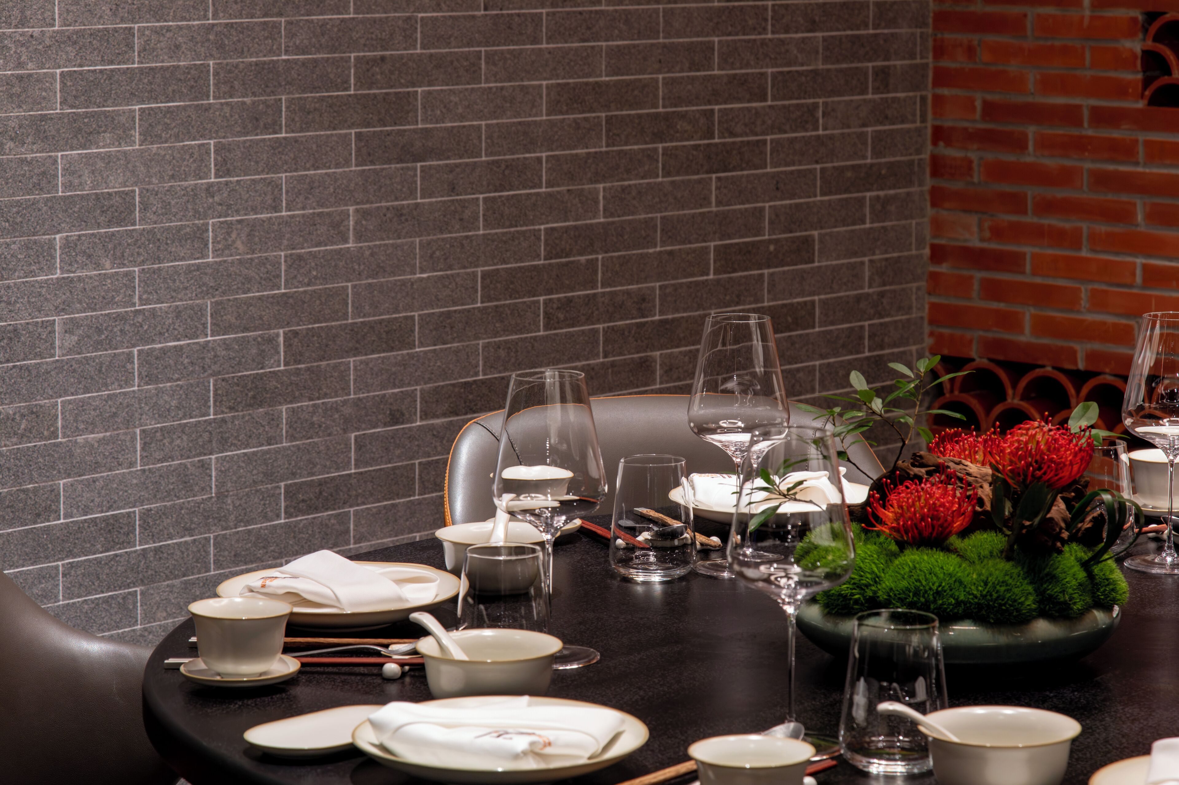 A dining table set with plates, bowls, and glasses, with a brick wall and a plant in the background.
