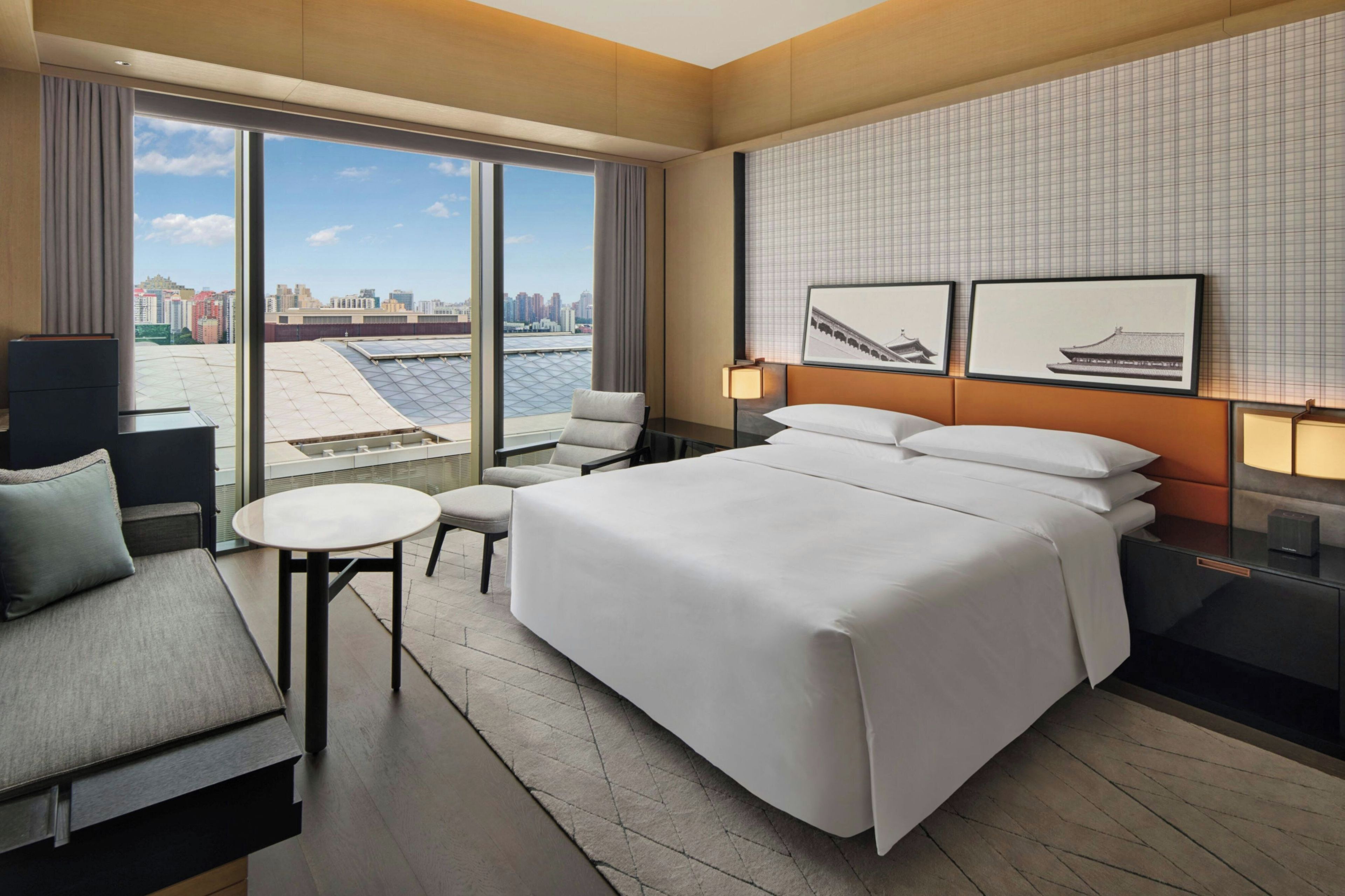 A hotel room with a bed, a desk, a chair, a small table, and a view of the cityscape through large windows.