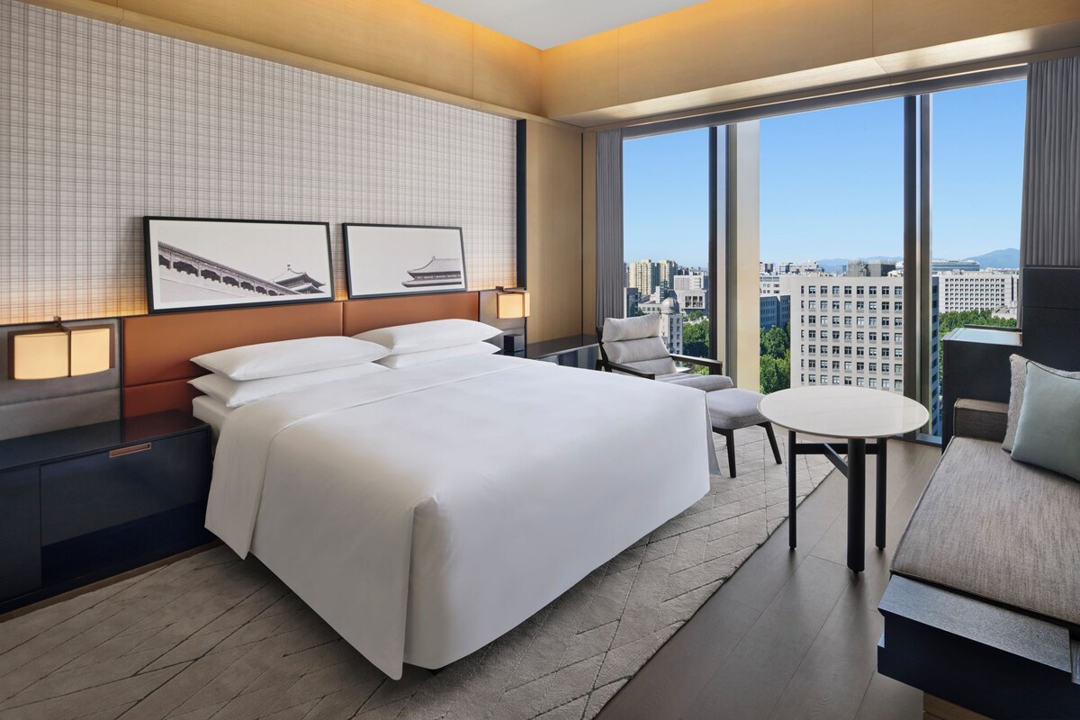 Una habitación de hotel moderna con una cama grande, un área de descanso con una mesa y vistas a la ciudad a través de amplias ventanas.