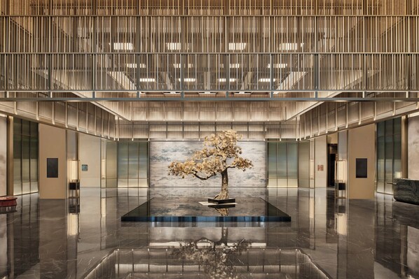 A spacious lobby with a central reflective floor, a large tree sculpture, and a modern design with vertical lines and glass partitions.
