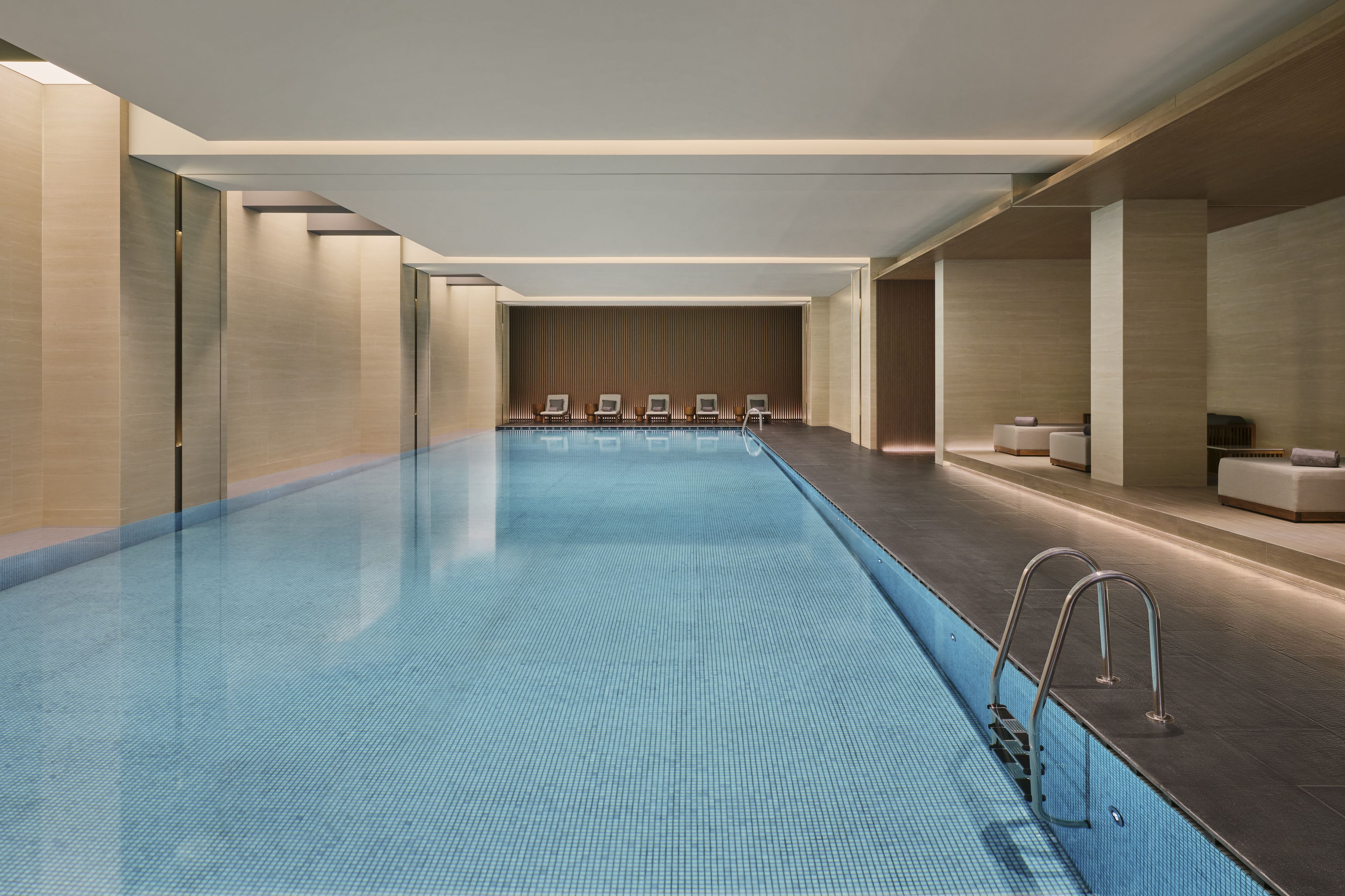 A modern indoor swimming pool with a clear blue tiled floor, surrounded by lounge chairs and a wooden wall with vertical slats.