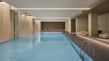 A modern indoor swimming pool with a clear blue tiled floor, surrounded by lounge chairs and a wooden wall with vertical slats.