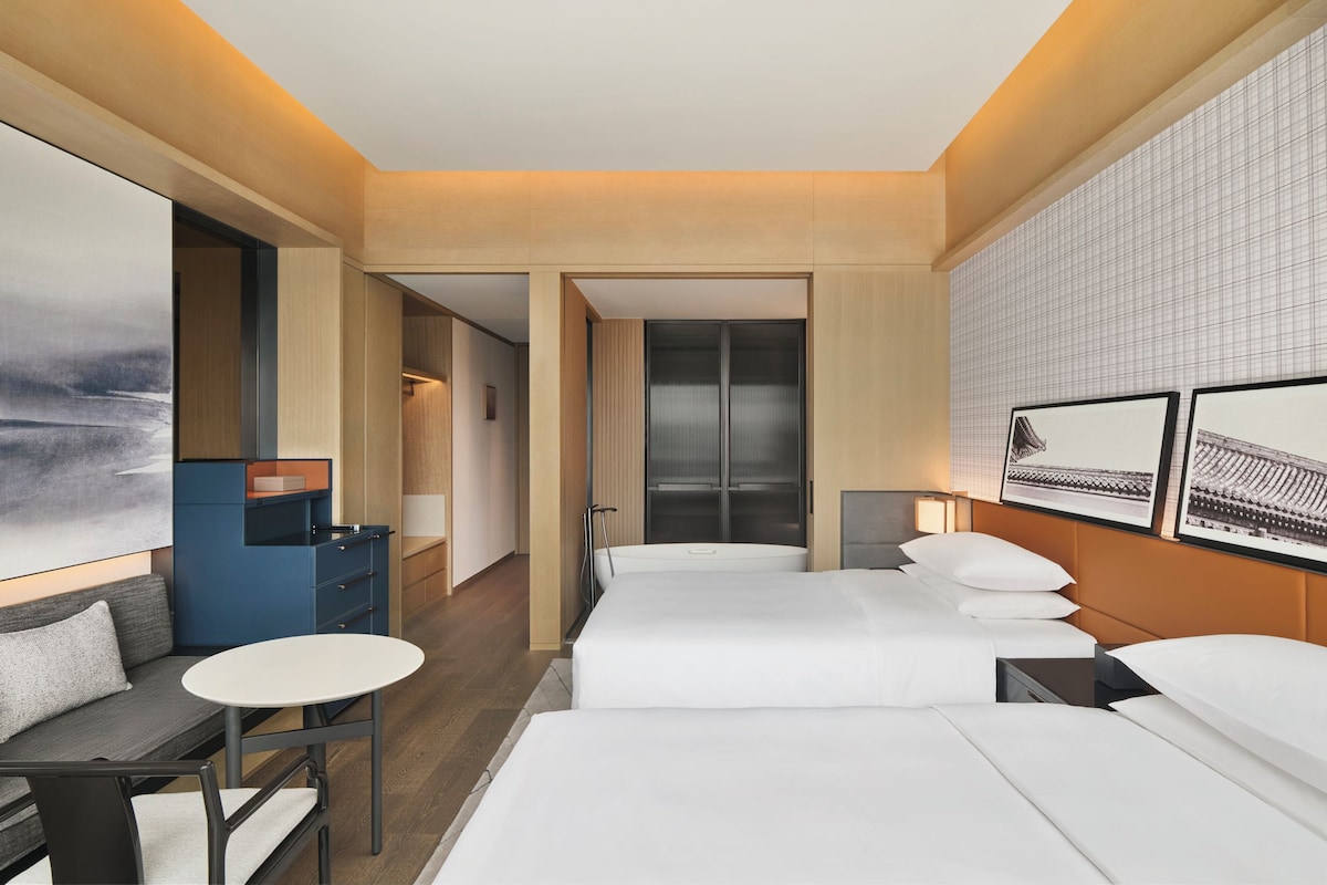 Quarto de hotel com cama, escrivaninha, cadeira e uma janela grande.