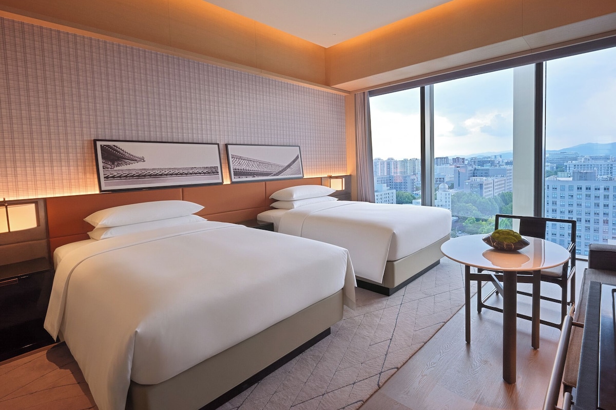 A hotel room with two beds, a small table, and a city view.