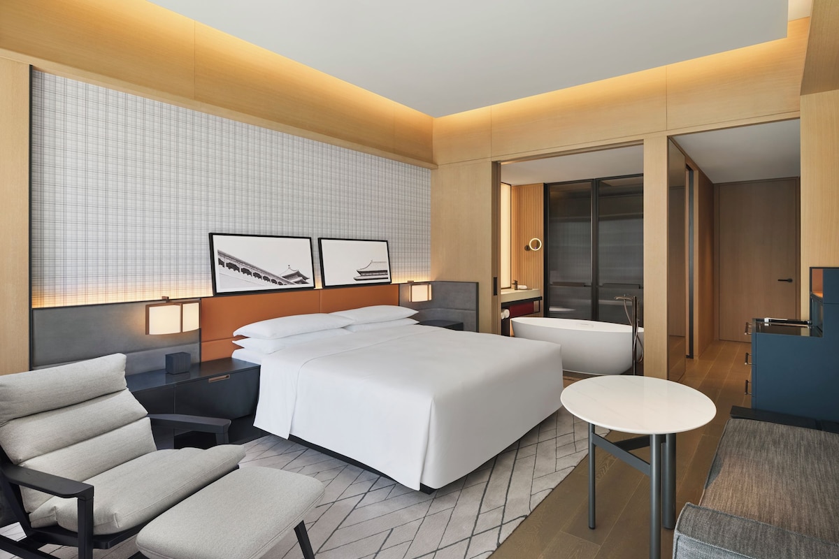 Quarto de hotel moderno com uma cama grande, uma escrivaninha, uma cadeira e uma mesa pequena.