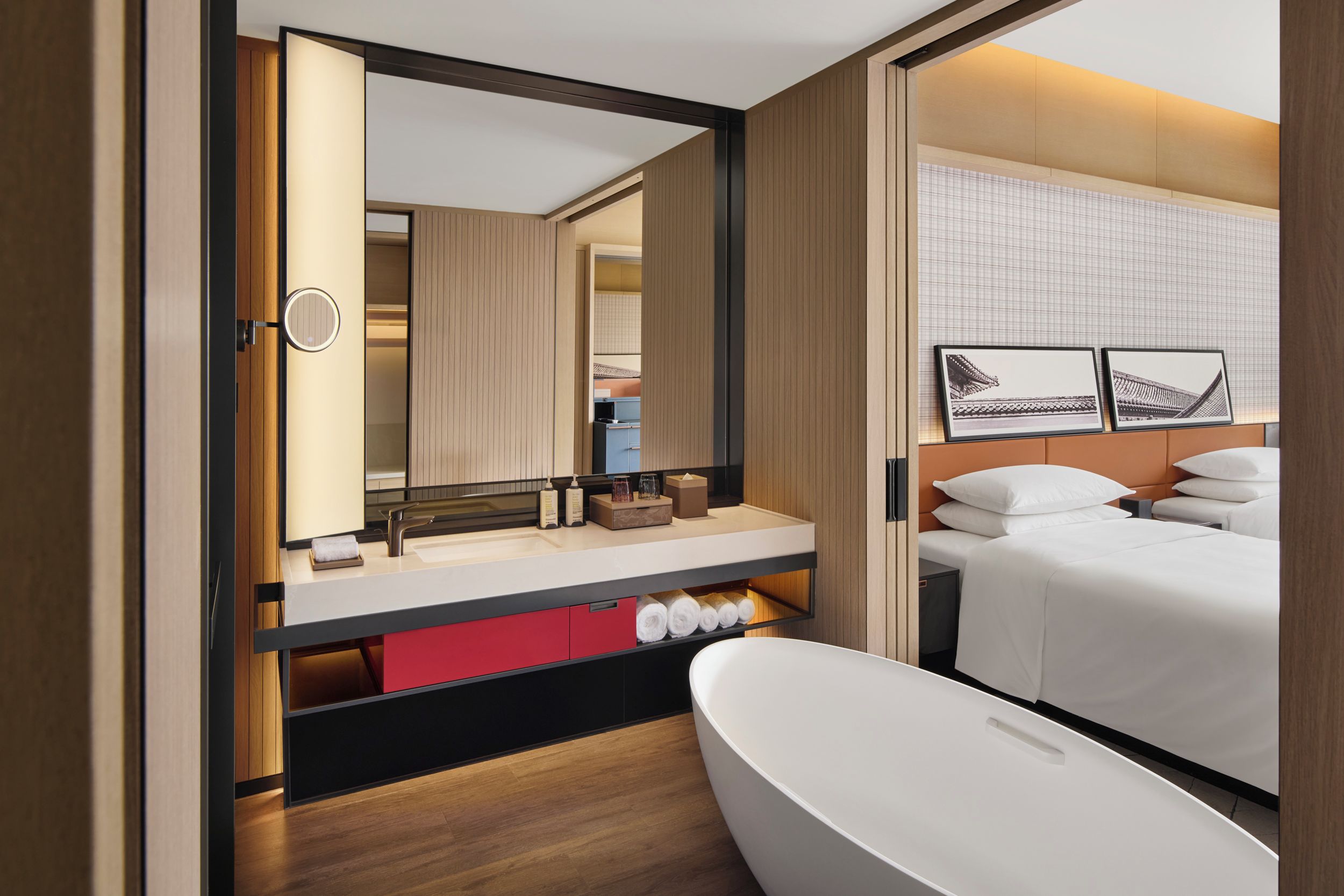 A modern hotel room with a large mirror, a bathtub, a bed with white linens, and a small bathroom area with a sink and toiletries.