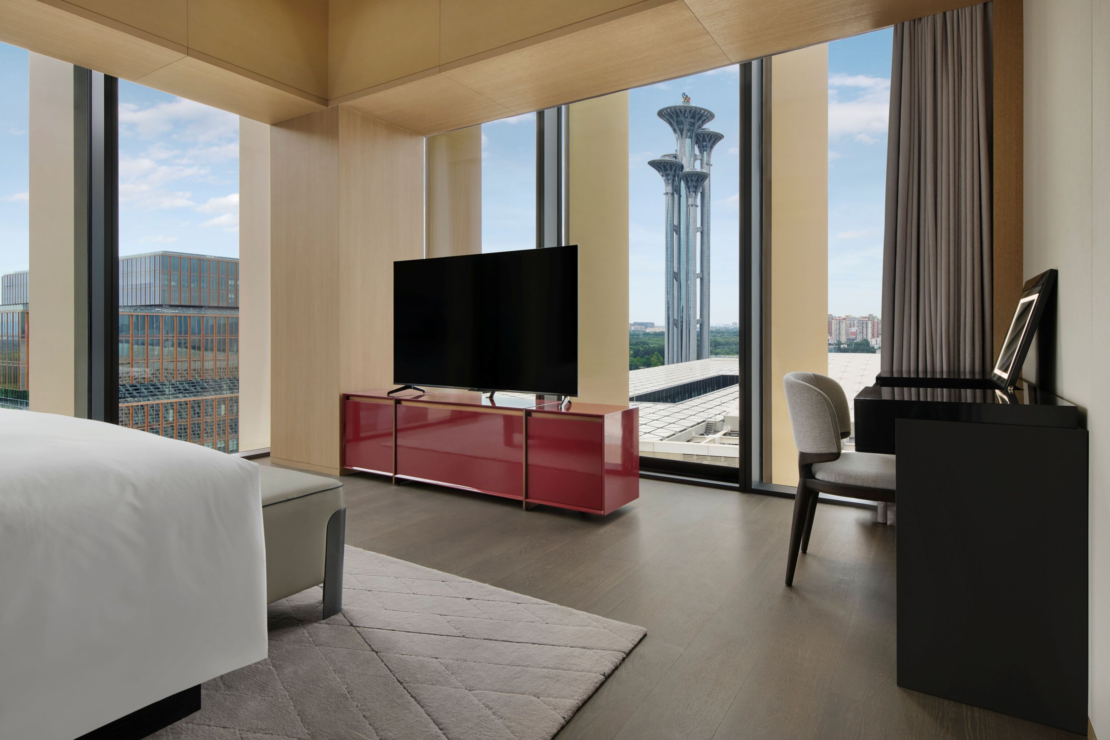 Une chambre d’hôtel moderne dotée d’un grand lit, d’un téléviseur à écran plat, d’un mini-bar rouge, d’un bureau avec une chaise et offrant une vue sur le paysage urbain et une tour.