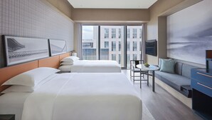 Habitación de hotel con dos camas, zona de estar, televisor y ventana grande con vista a la ciudad.