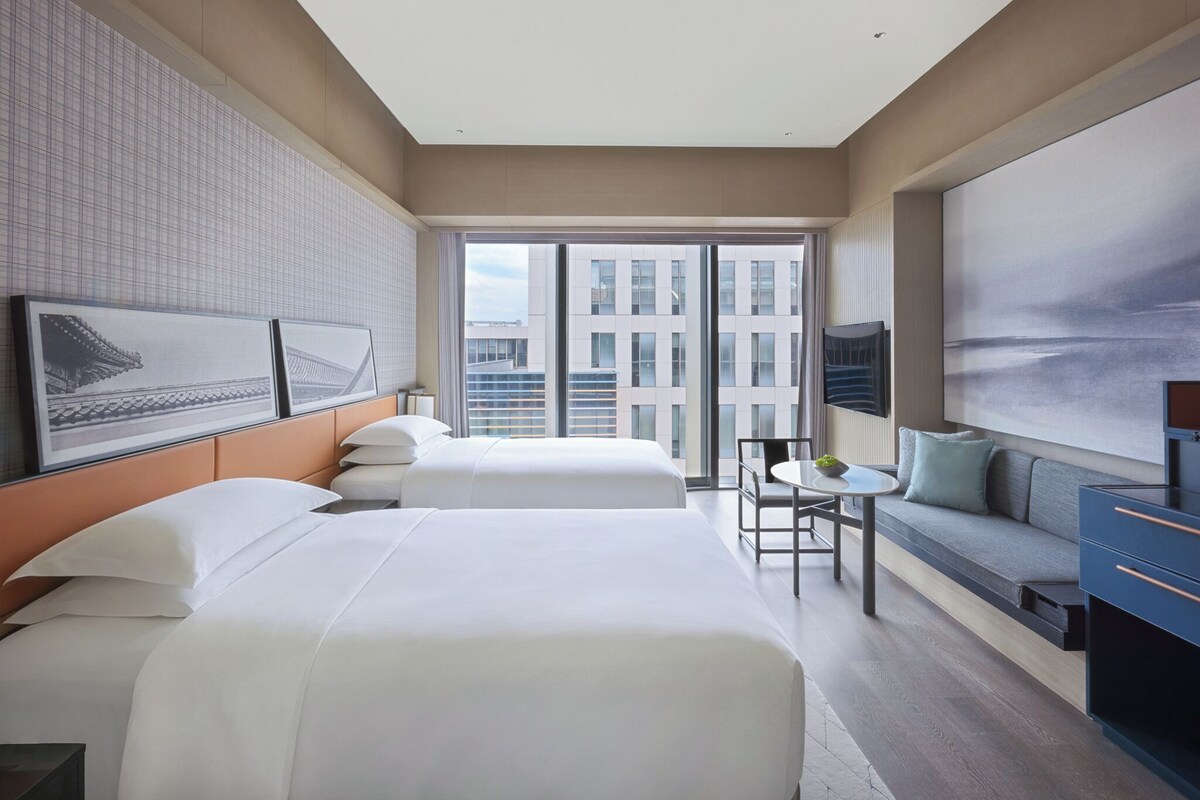 A hotel room with two beds, a seating area, a TV, and a large window with a city view.