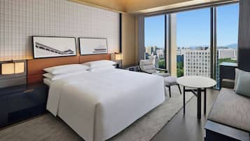 Habitación de hotel con una cama grande, un escritorio, una silla y una mesita. Hay dos cuadros en la pared, y la habitación tiene ventanas amplias con vista a la ciudad.
